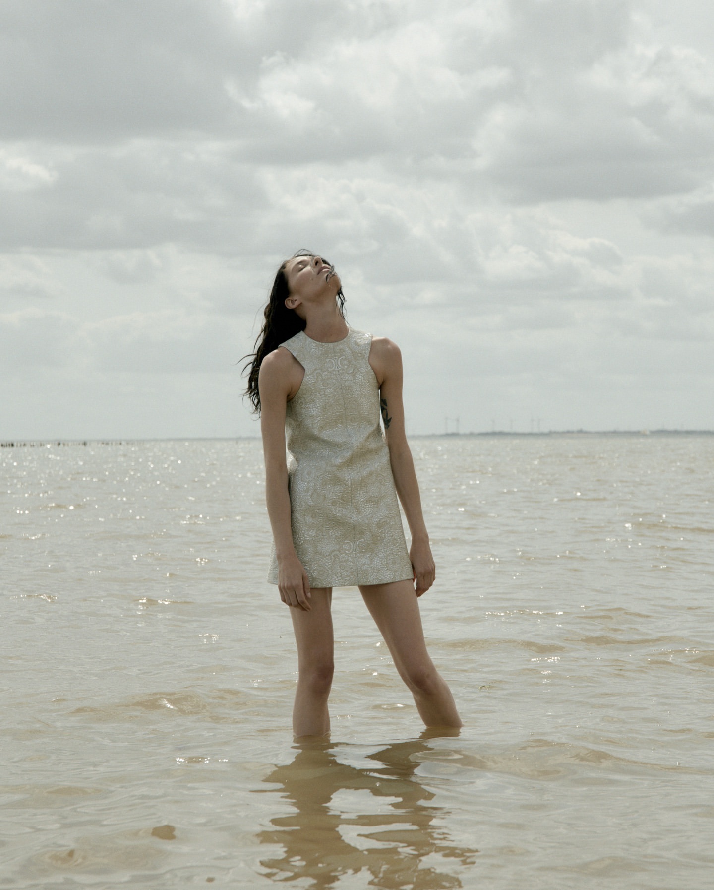 An adult female stands in shallow water wearing a patterned mini dress with her head tilted back looking towards the sky on a cloudy day.