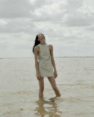 An adult female stands in shallow water wearing a patterned mini dress with her head tilted back looking towards the sky on a cloudy day.