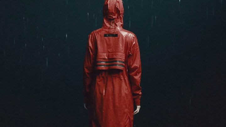 Rear view of an adult wearing a red hooded raincoat in the rain. The background is a dark, solid colour with rain streaks.