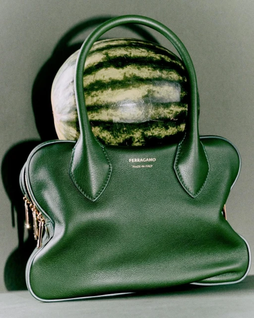 A green leather purse sits beneath a watermelon in a studio shot with a grey background and soft lighting.