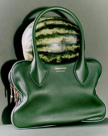 Green leather handbag with a whole watermelon placed inside the open top against a dark studio backdrop