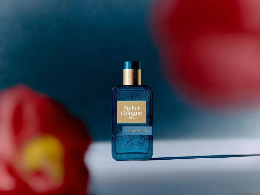 Turquoise Atelier Cologne bottle on a white surface with red flower-shaped lights against a blue background in a studio setting.