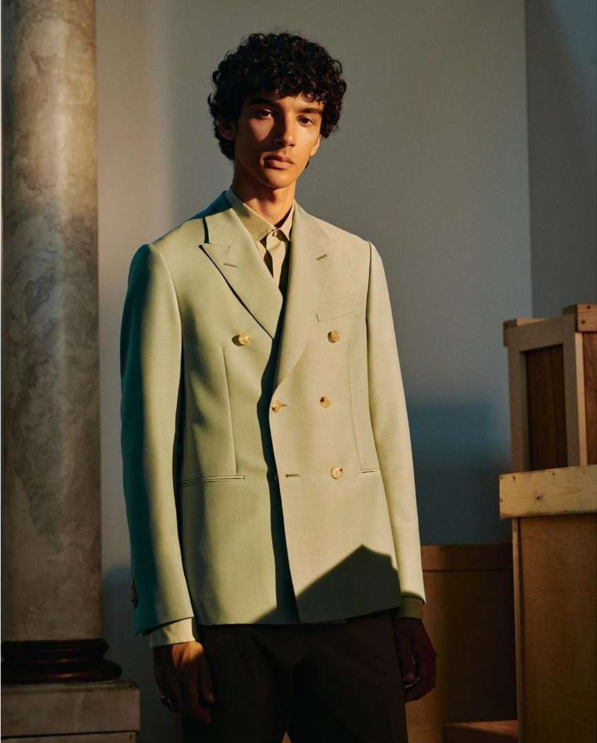 Adult male poses indoors wearing a light green double-breasted blazer, brown pants, and a button-down shirt near a decorative column.