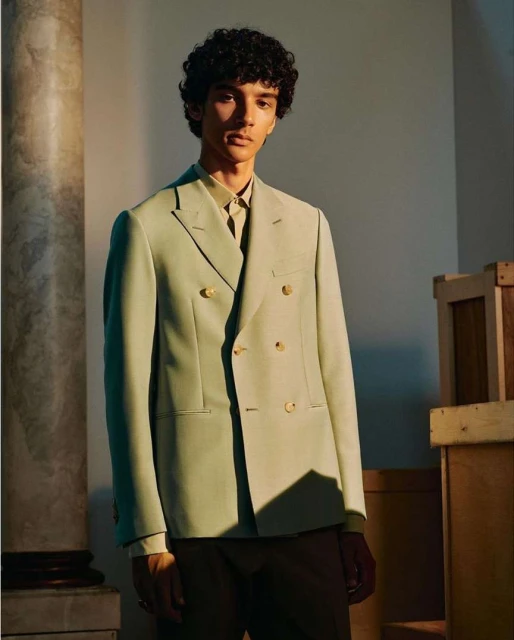 Adult male poses indoors wearing a light green double-breasted blazer, brown pants, and a button-down shirt near a decorative column.