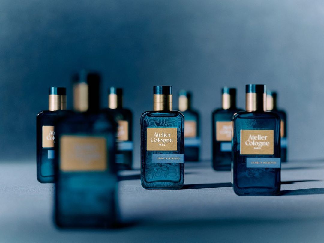 Several blue Atelier Cologne perfume bottles with gold caps and labels are arranged against a soft blue background. Studio shot.