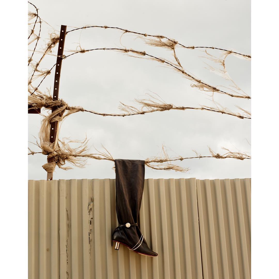Fashionable knee-high boot posed on a corrugated metal fence with a barbed wire accent against an overcast sky backdrop.