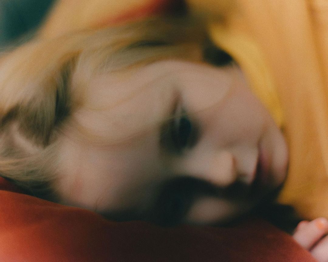 Close-up of child's face with blond hair, red pillow, and yellow fabric in a blurred, soft-focus image with warm, diffused lighting.