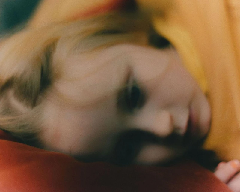 Close-up of child's face with blond hair, red pillow, and yellow fabric in a blurred, soft-focus image with warm, diffused lighting.