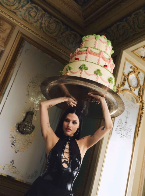 A woman in a black dress stands indoors holding a large three-tiered cake on a silver platter, set against an ornate, gilded background.