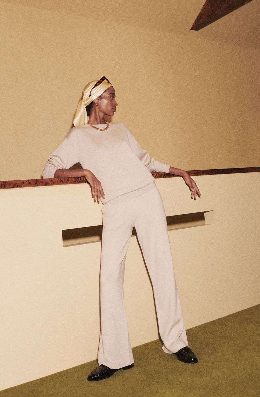 Model in cream knit top and trousers wearing a yellow headscarf leaning against a beige wall by photographer Joyce Ng.