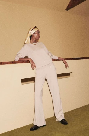 Model in cream knit top and trousers wearing a yellow headscarf leaning against a beige wall by photographer Joyce Ng.