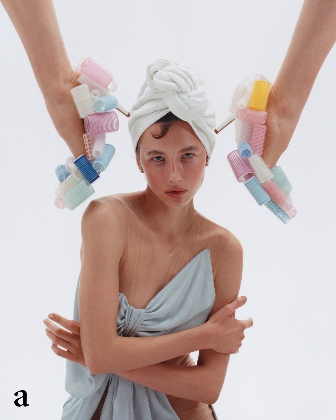Elegant woman with head towel and blue dress, surrounded by hands holding curlers against a bright white background in a beauty editorial.