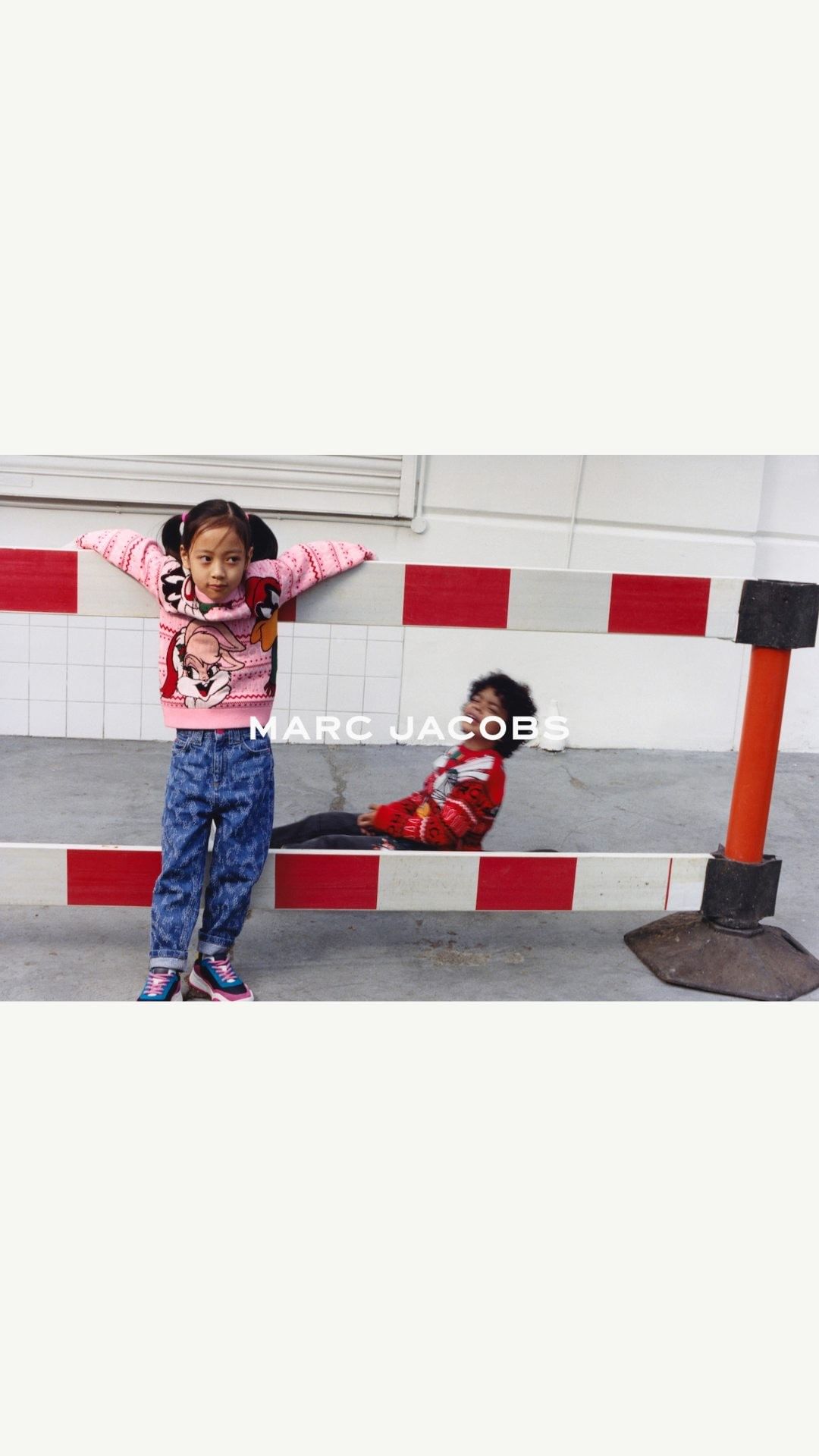 Two children pose near a red and white barrier, one standing with arms out, the other reclining. The clothing features designs and the brand logo.