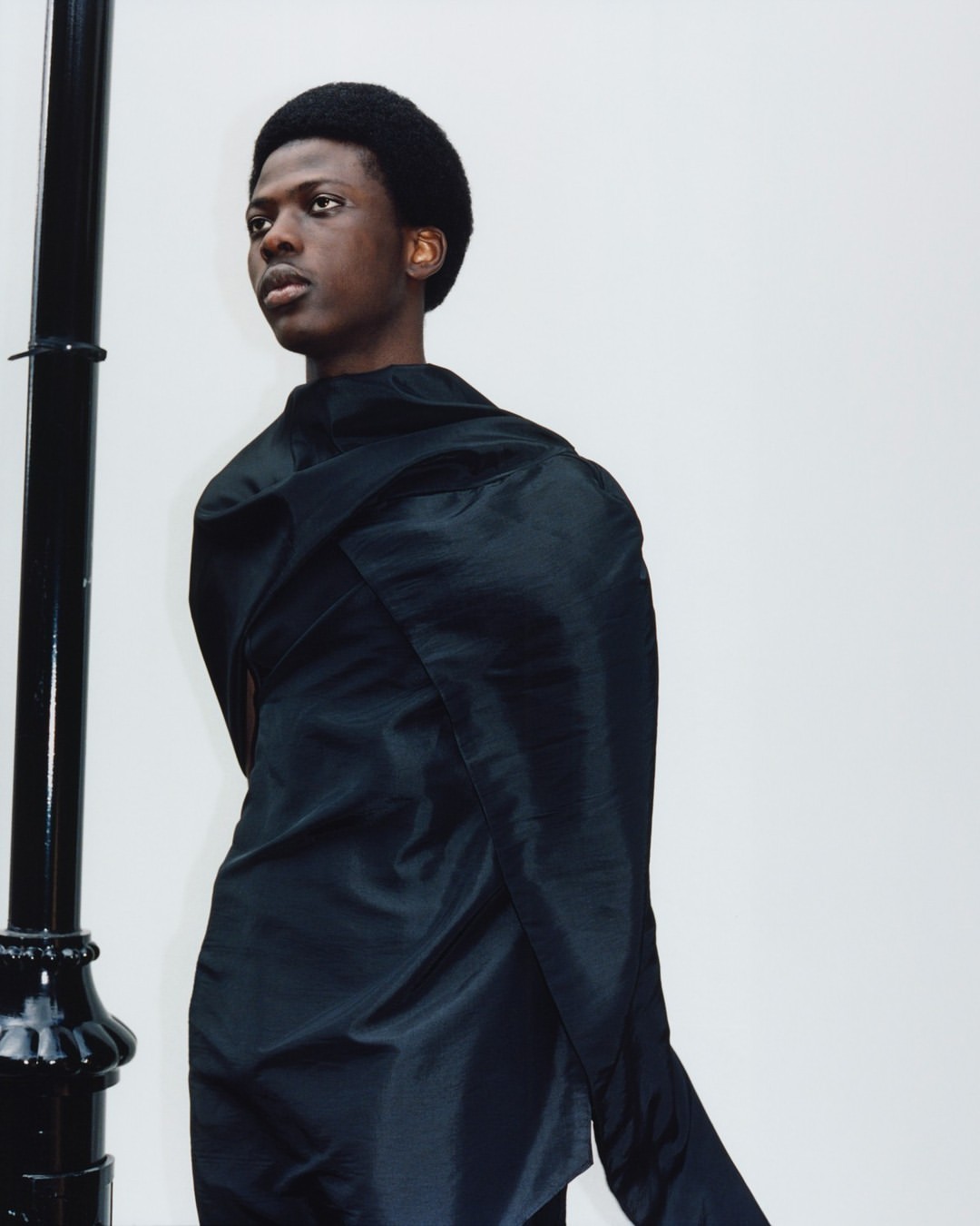Portrait of an African-American male model in a dramatic black draped garment standing near a black lamp post against a white wall.