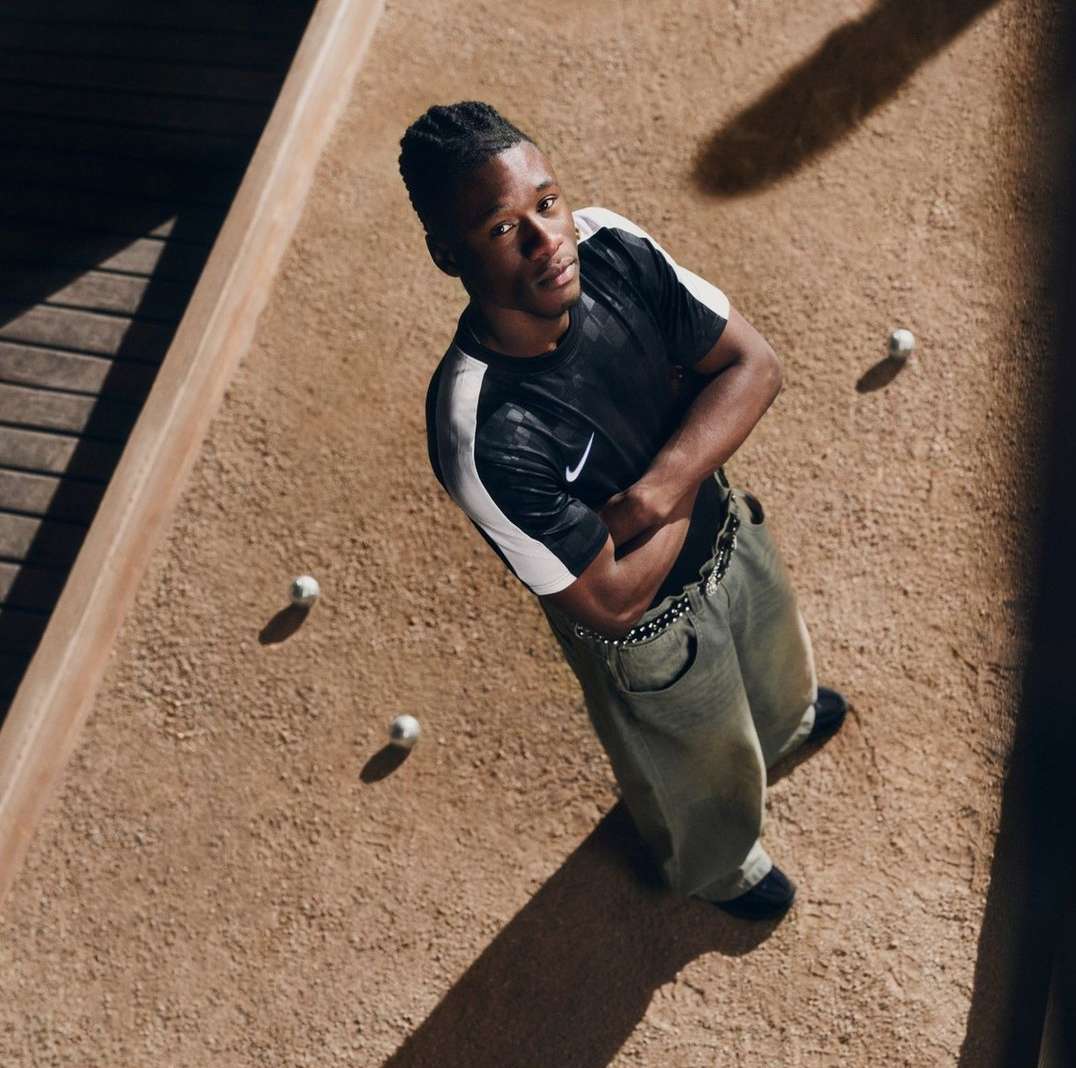 An adult male stands in a low-angle full shot on a bocce court with a Nike shirt, baggy jeans, and dark sneakers, looking at the camera.