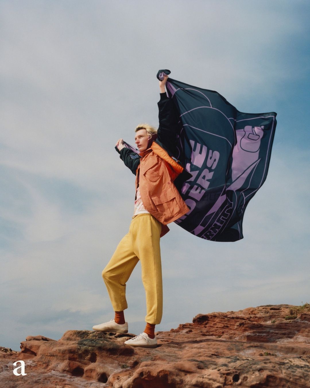 Man holding a branded flag on a rocky outcrop against a cloudy sky. He wears a jacket, t-shirt, trousers, and shoes, standing outside.