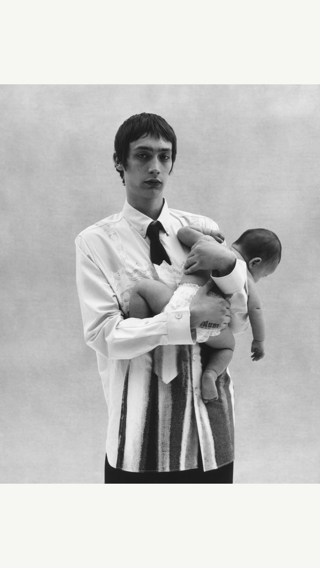 Black-and-white studio shot of a young adult male holding a baby, both looking at the camera, in a vertical composition with neutral backdrop.