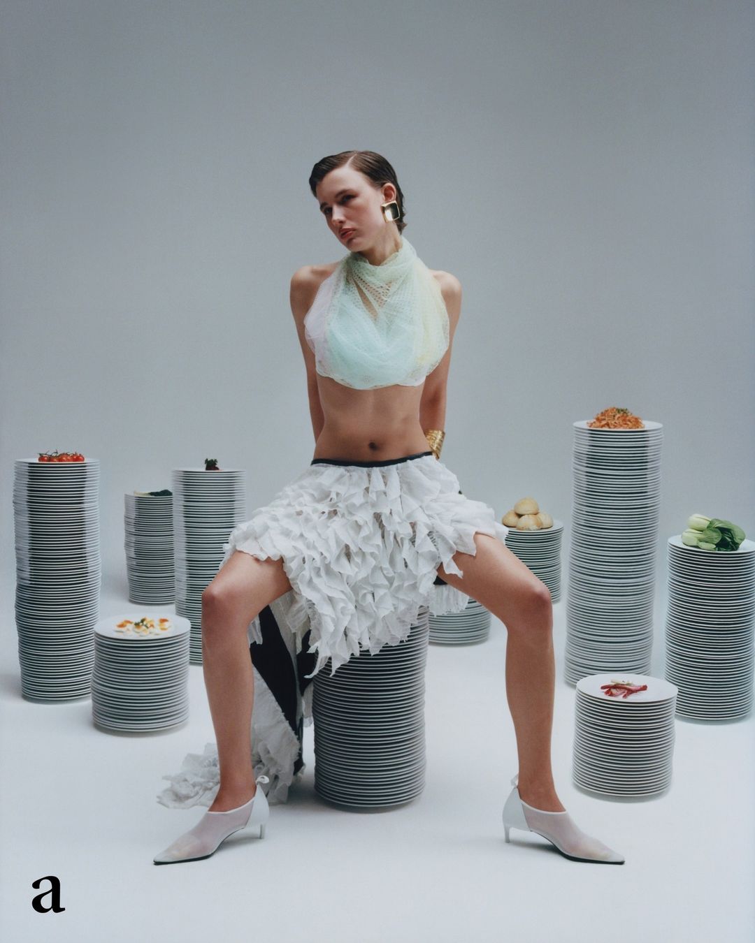 Fashionable woman poses in white tulle skirt and halter top, seated atop stack of plates surrounded by stacks of plates.
