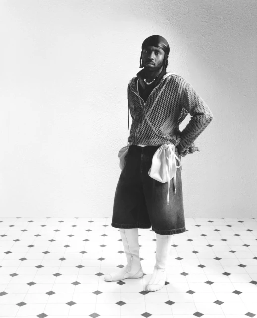 Black and white shot of a Black male model in streetwear fashion posing in a light studio with a tiled floor and plain white walls.