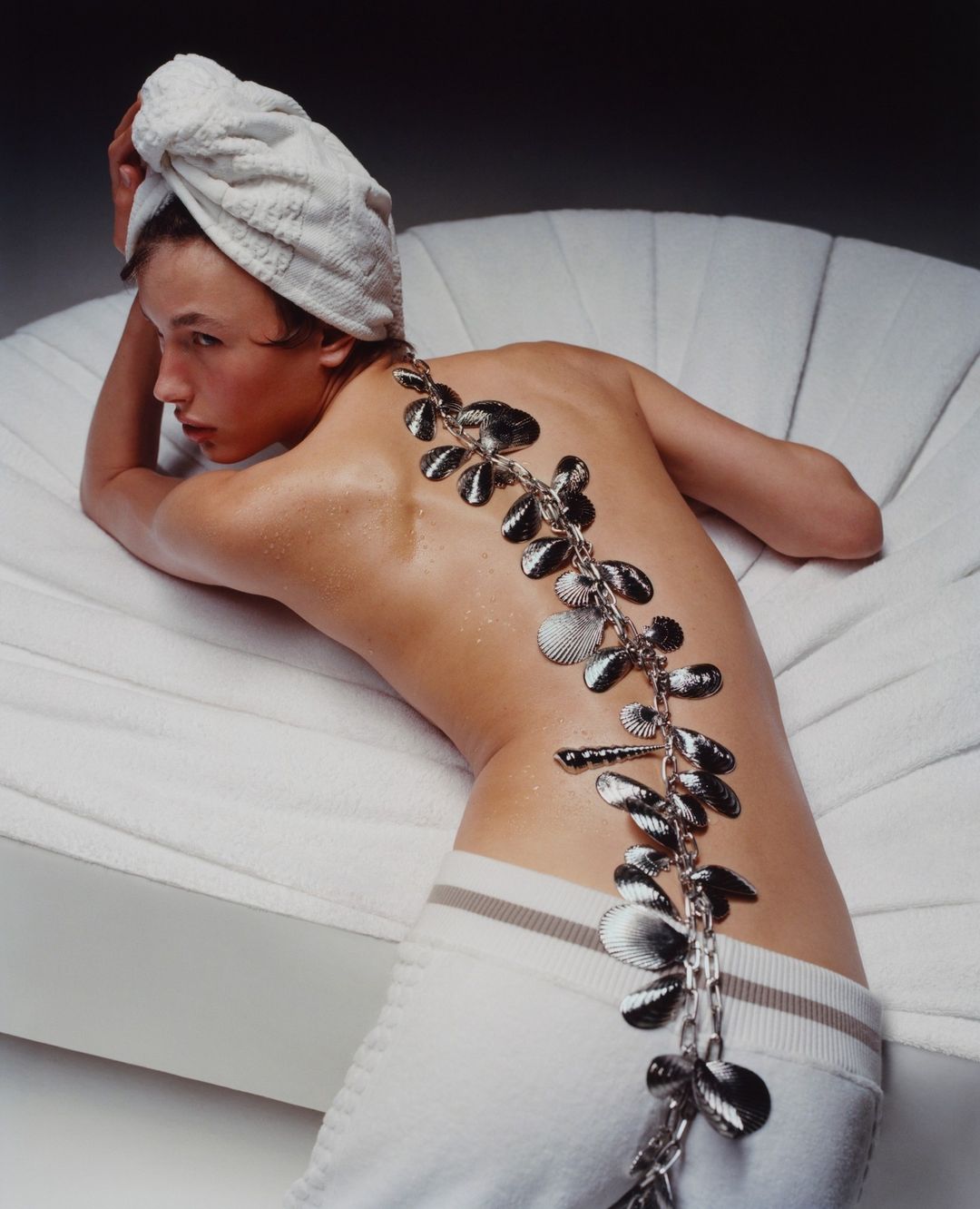 Overhead shot of a person lying on a white sofa with a towel on their head and a decorative shell necklace.