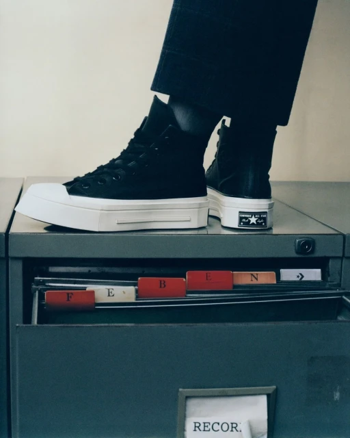Elevated black high-top sneakers with thick white platform soles, displayed on a metal filing cabinet in a brightly lit indoor space.