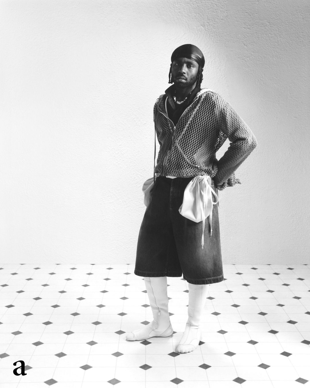 Black male model in knit zip jacket, denim shorts, and white boots poses in a studio with white walls and a patterned floor.