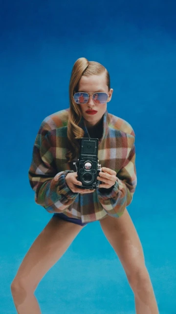 Woman with retro camera in a studio. She's wearing a plaid jacket, sunglasses, and has blonde hair. The backdrop is a bright blue.