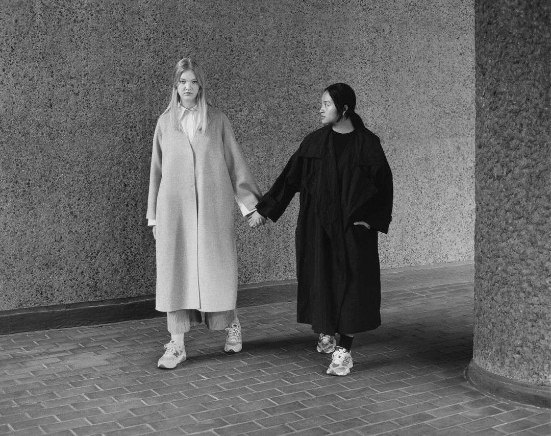 Two adult women in coats walk hand-in-hand against a mottled grey wall on a brick walkway in this black and white lifestyle shot.