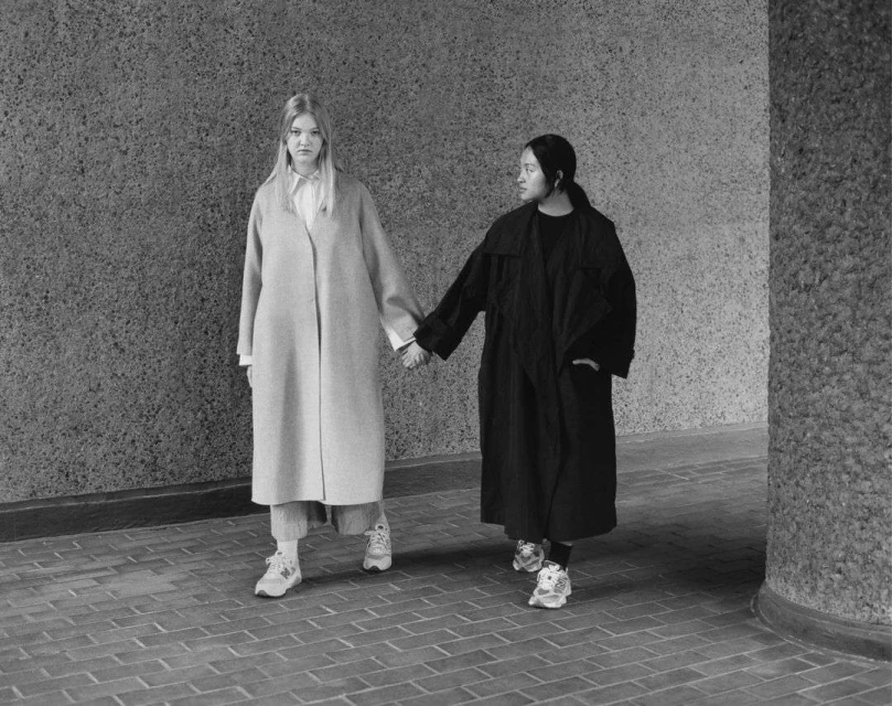 Two adult women in coats walk hand-in-hand against a mottled grey wall on a brick walkway in this black and white lifestyle shot.
