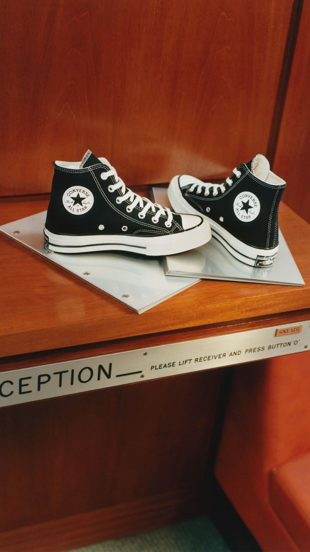Two Converse Chuck Taylor All-Star high-top sneakers, one positioned to the side, on a wooden surface with a silver plate.