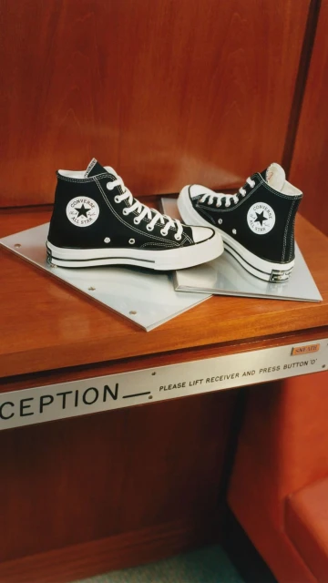 Two Converse Chuck Taylor All-Star high-top sneakers, one positioned to the side, on a wooden surface with a silver plate.