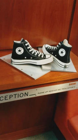 Two Converse Chuck Taylor All-Star high-top sneakers, one positioned to the side, on a wooden surface with a silver plate.