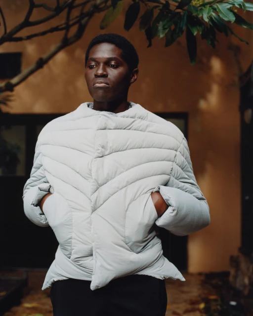 An adult male model with dark skin, wearing a light grey puffer jacket stands outdoors in front of a brown building, surrounded by foliage.