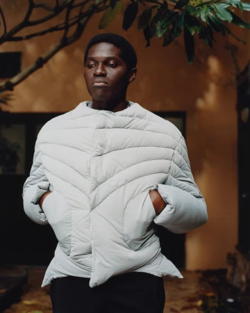An adult male model with dark skin, wearing a light grey puffer jacket stands outdoors in front of a brown building, surrounded by foliage.
