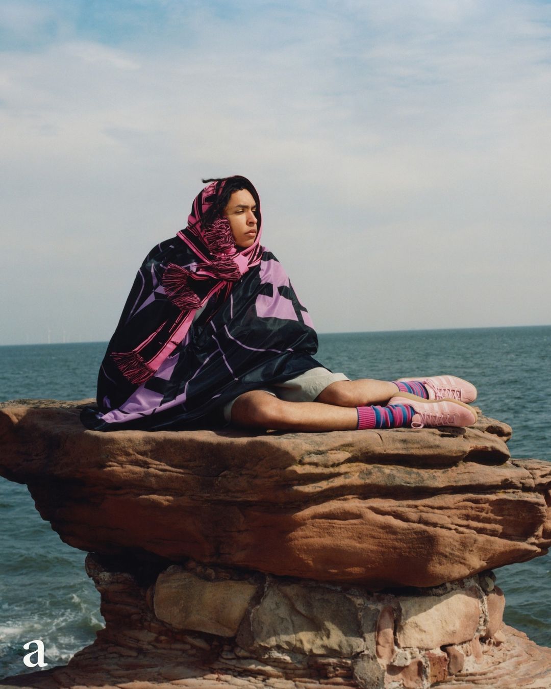 A person wrapped in a dark patterned blanket and pink scarf seated on a rocky outcrop by the ocean under an overcast sky.