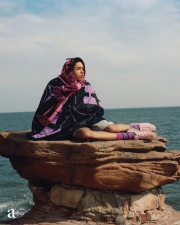 A person wrapped in a dark patterned blanket and pink scarf seated on a rocky outcrop by the ocean under an overcast sky.