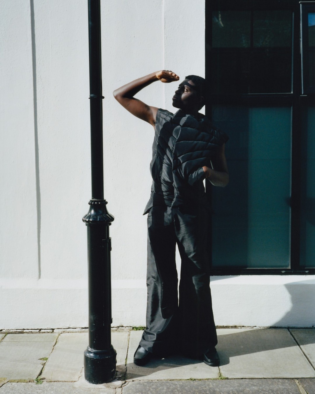 Young Black man shielding his eyes in sunlight, wearing a puffy vest and wide-leg pants, stands next to a vintage-style black lamppost outdoors.