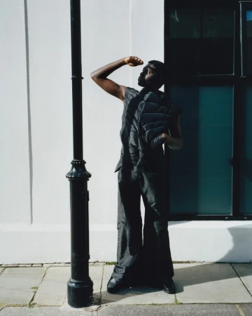 Young Black man shielding his eyes in sunlight, wearing a puffy vest and wide-leg pants, stands next to a vintage-style black lamppost outdoors.