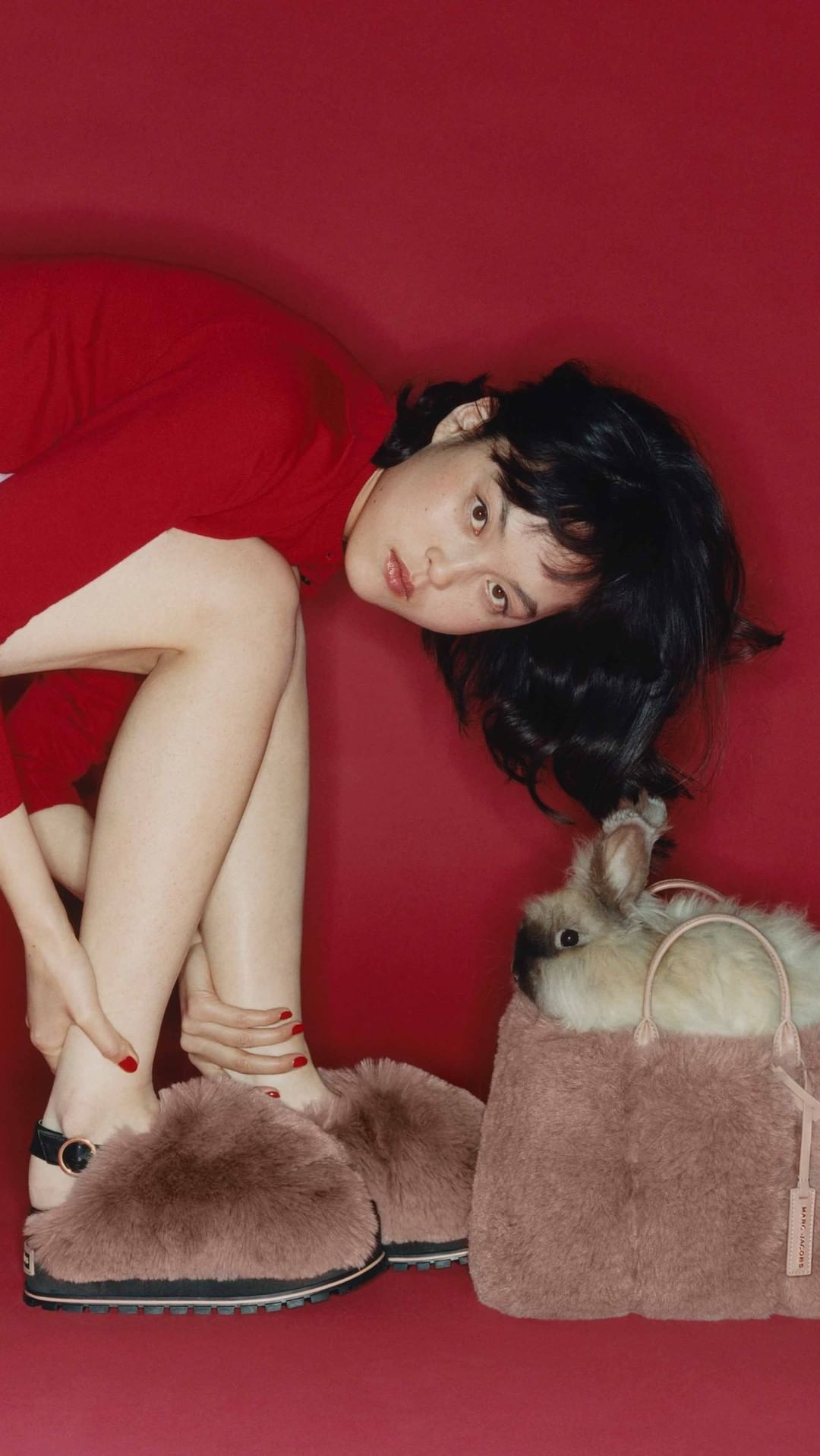 Woman wearing red dress and furry slippers, next to a fluffy pink bag containing a rabbit, against a red studio backdrop. Fashion shot.