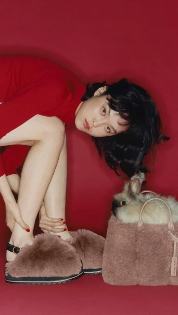 Woman wearing red dress and furry slippers, next to a fluffy pink bag containing a rabbit, against a red studio backdrop. Fashion shot.