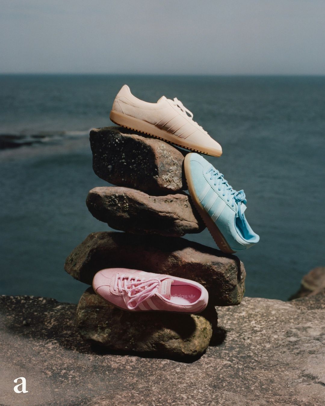 Three Adidas Gazelle sneakers in beige, light blue, and pink stacked on rocks outdoors with an ocean background, fashion product shot.