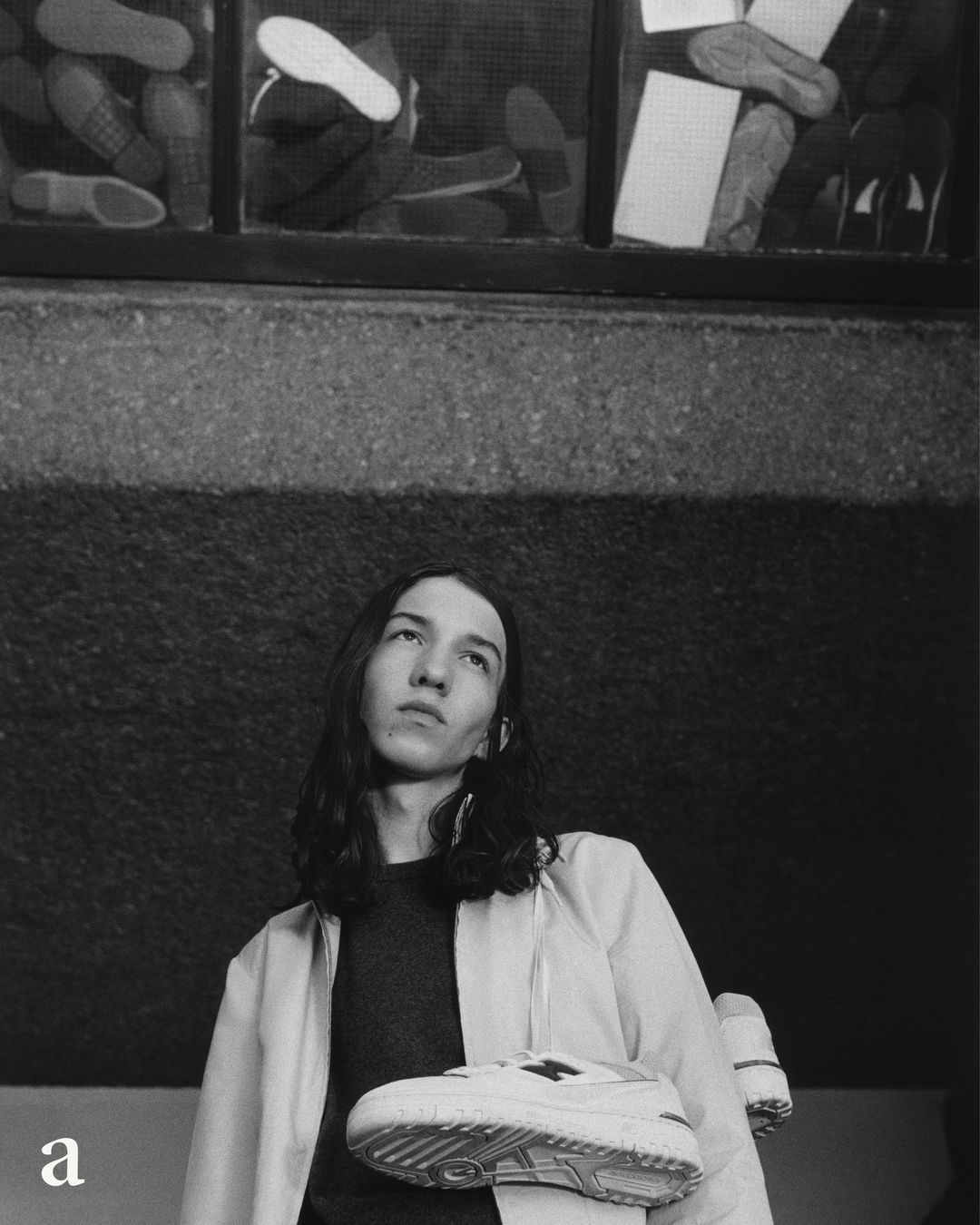 Monochrome fashion portrait of a young adult with long dark hair holding up a white athletic shoe against a textured wall.