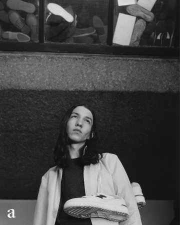 Monochrome fashion portrait of a young adult with long dark hair holding up a white athletic shoe against a textured wall.