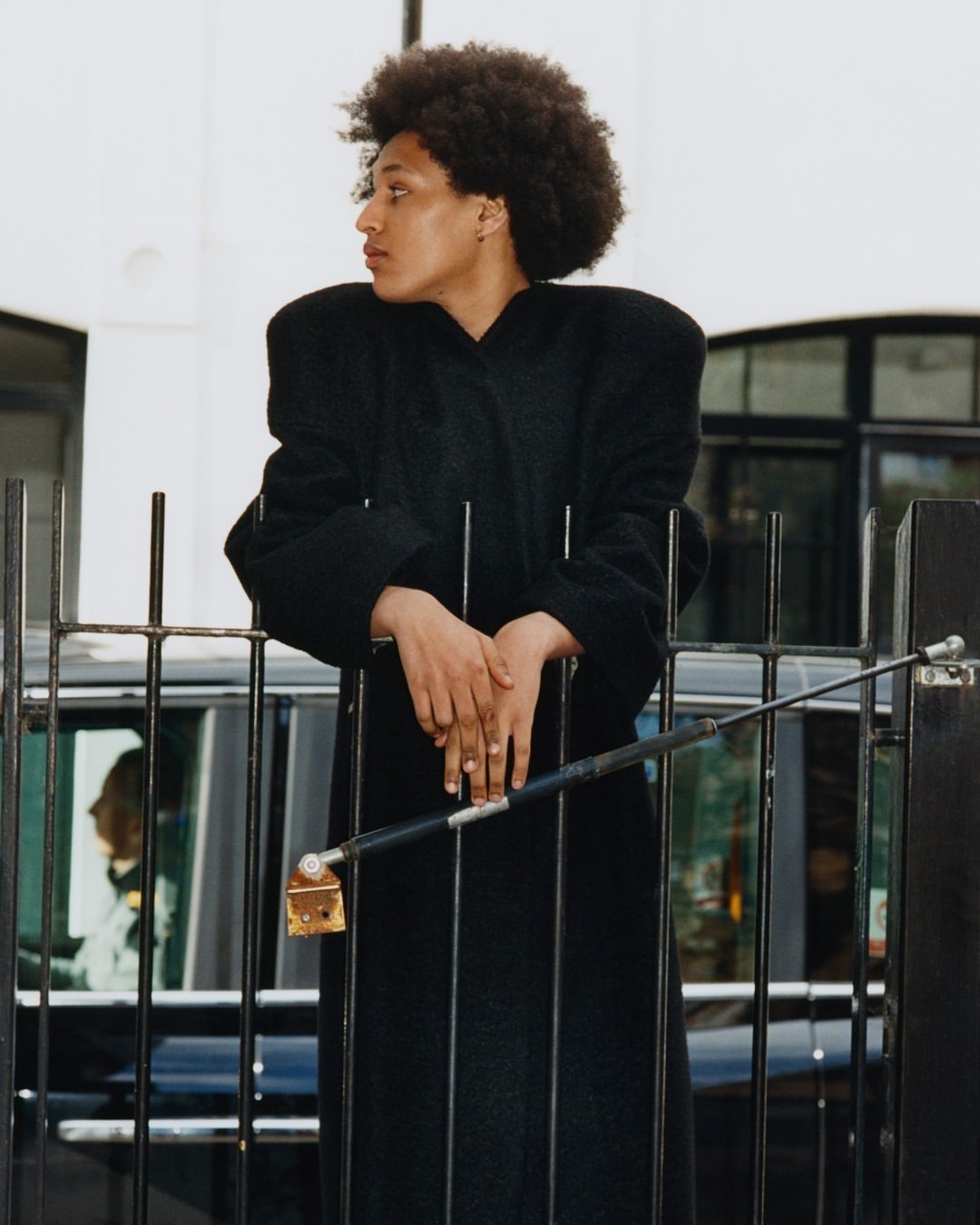 An elegant Black woman in a long black coat leans against a metal fence with a black car in the background, showcasing a fashion editorial shot.