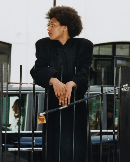 An elegant Black woman in a long black coat leans against a metal fence with a black car in the background, showcasing a fashion editorial shot.