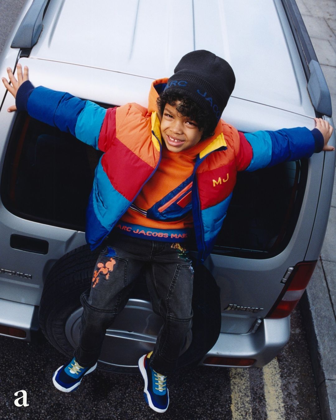 Young boy in a colorful puffer jacket sits on the back of a silver car, showcasing a casual, fashionable look on an urban street.