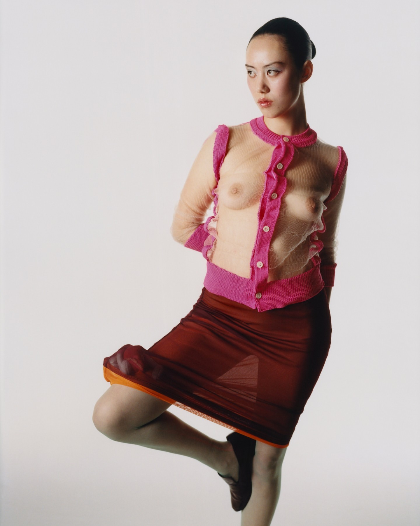 Adult woman poses in a sheer pink cardigan and red skirt in a studio against a white background in a fashion photograph.
