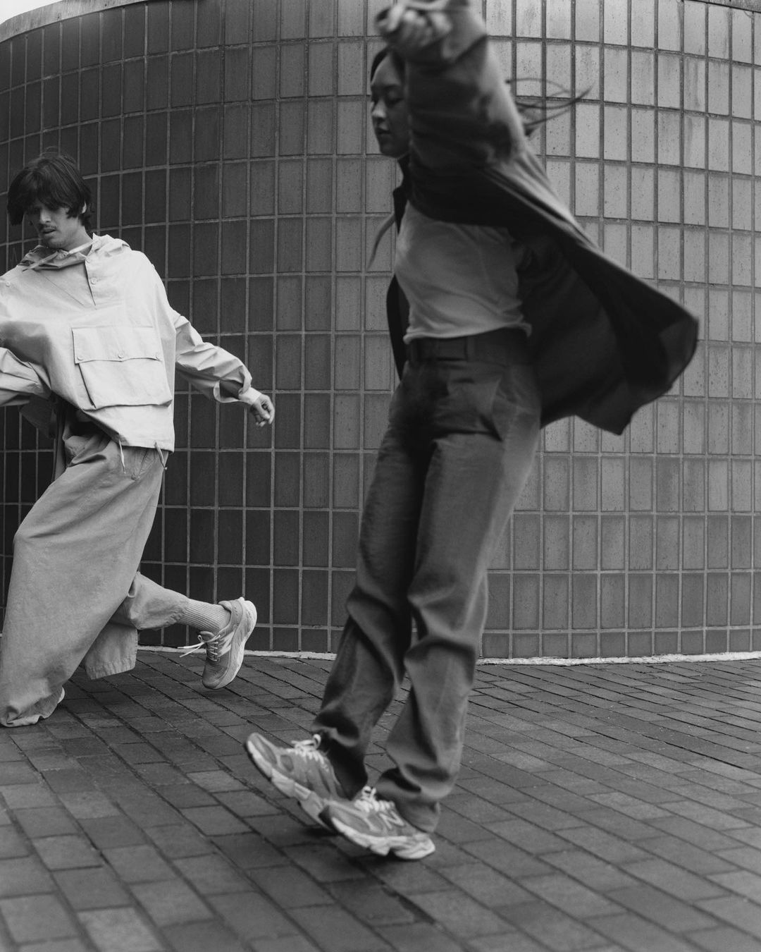 Two people in motion. One adult female wears a jacket, t-shirt, and jeans. One adult male wears a sweatshirt and pants on a paved surface.
