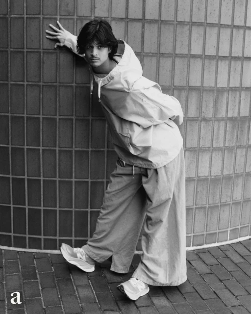 Young adult male fashion model poses outdoors in a hooded jacket and wide-leg pants, leaning against a tiled wall with running shoes.