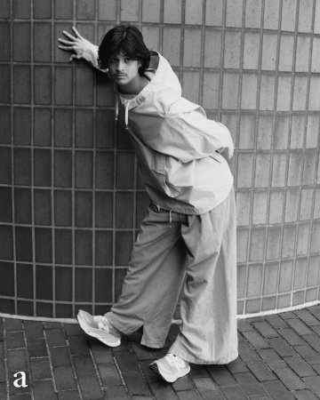 Young adult male fashion model poses outdoors in a hooded jacket and wide-leg pants, leaning against a tiled wall with running shoes.